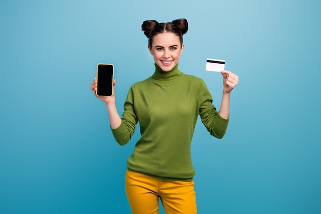 Foto de dama alegre mantenga tarjeta de crédito nuevo modelo de teléfono inteligente que muestra el servicio de presentación de pago en línea usar pantalones amarillos de cuello alto verde pared de color azul aislado