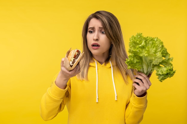 Foto de dama atractiva elige entre hamburguesa y ensalada, comida sana o chatarra. Viste con capucha amarilla casual, fondo de color amarillo aislado.