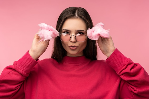 Foto de dama atractiva en gafas juega con dulce de algodón de azúcar se divierte feliz usar suéter rosa casual aislado fondo de color rosa