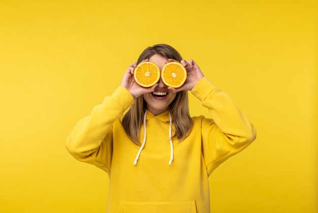 Foto de dama atractiva sostiene naranjas en rodajas y cubre los ojos con una sonrisa. Viste con capucha amarilla casual, fondo de color amarillo aislado.