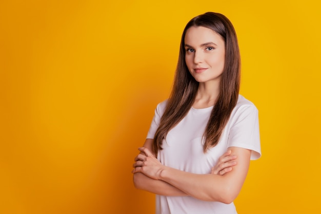 Foto de dama segura de sí misma con los brazos cruzados, mira la cámara, usa camiseta blanca posando sobre fondo amarillo