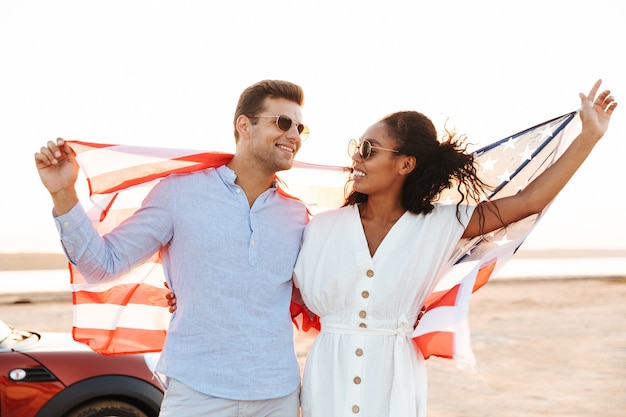 Foto de alegre casal multiétnico, homem e mulher, sorrindo e segurando a bandeira americana ao ar livre