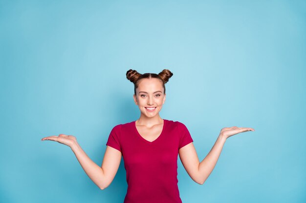 Foto de alegre fofa doce muito charmosa garota segurando dois objetos com as mãos, mostrando os dois lados de um problema a ser resolvido vestindo uma camiseta vermelha isolada sobre uma parede azul pastel