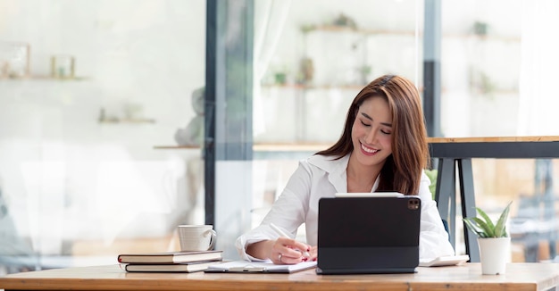 Foto de alegre mulher asiática bonita, tendo sido empregada para trabalhar como executivo, sorrindo dentuçosamente, sentado na área de trabalho com o laptop e escrevendo nota.