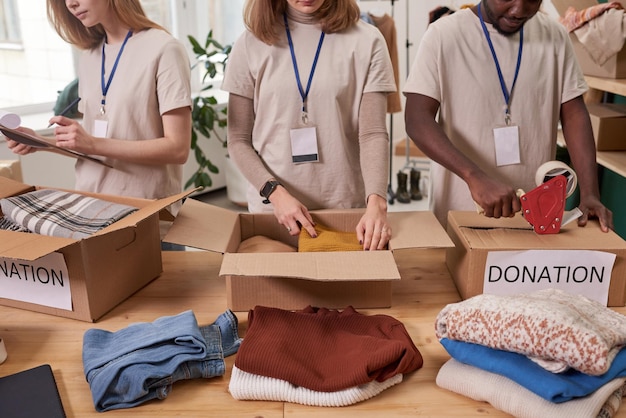 Foto foto de alto ângulo de três voluntários irreconhecíveis vestindo camisetas bege embalando roupas em caixas de papelão para doação