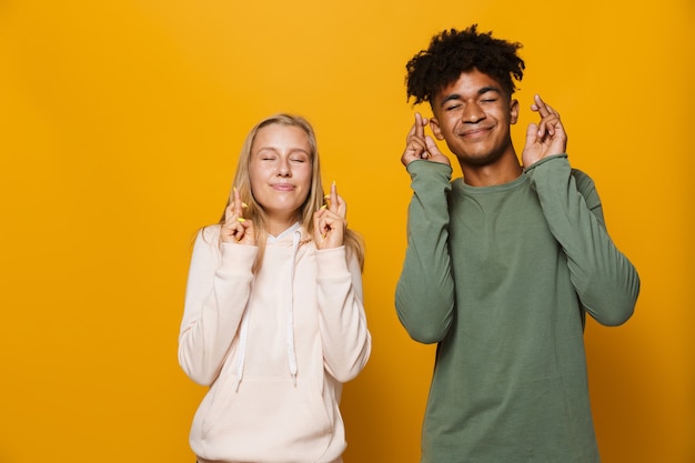 Foto de amigos adolescentes, homem e mulher, de 16 a 18 anos, mantendo os dedos cruzados e sonhando com boa sorte, isolado sobre um fundo amarelo