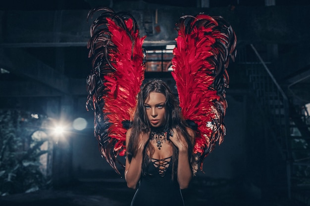 Foto de arte de uma mulher bonita de Dark Angel. Uma garota com asas de diabo vermelho e um vestido preto no quarto escuro