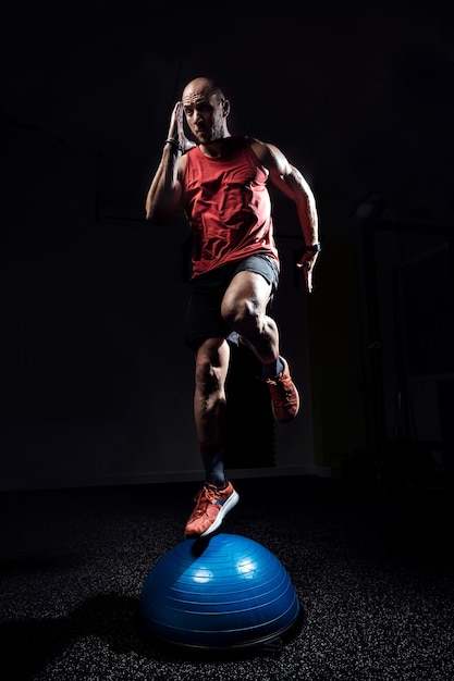 Foto de atleta apto realizando exercícios na bola de bosu do hemisfério de ginástica no estúdio escuro.