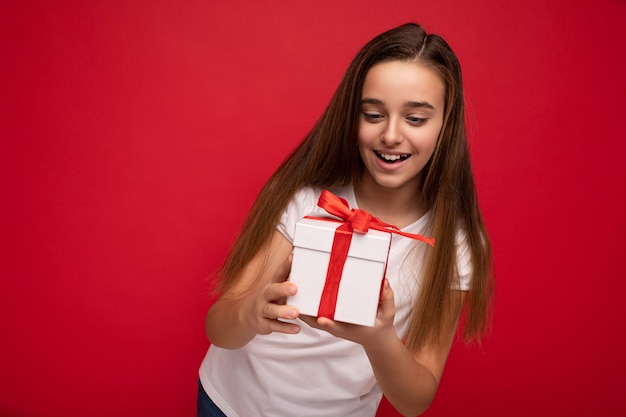 Foto de atraente menina morena sorridente feliz isolada sobre a parede de fundo vermelho, vestindo uma camiseta branca, segurando uma caixa de presente branca com fita vermelha e olhando o presente.