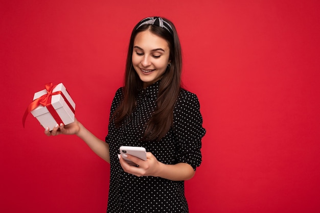 Foto de atraente menina morena sorridente positiva fofa isolada sobre a parede de fundo vermelho, vestindo uma blusa preta, segurando uma caixa de presente branca com fita vermelha e olhando para a tela do telefone móvel.