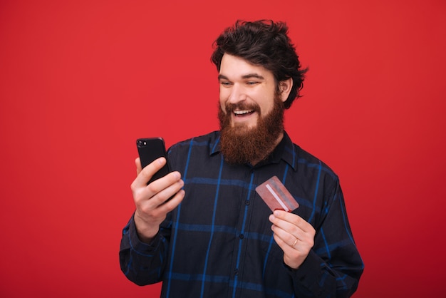 Foto de barbudo, usando smartphone e segurando um novo cartão de crédito sobre parede vermelha