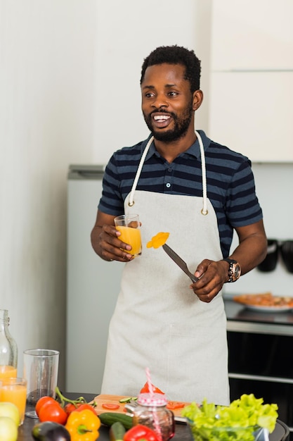 Foto de belo jovem de pele escura em óculos segurando um copo com suco de laranja espremido na hora, desfrutando de uma bebida saborosa na cozinha leve em casa pela manhã, desviando o olhar com expressão satisfeita.