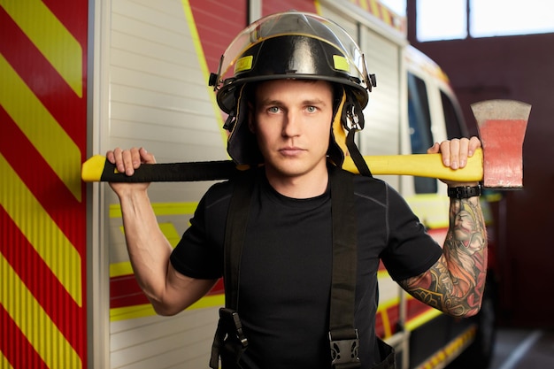 Foto foto de bombeiro usando capacete com machado contra carro de bombeiros imagem de jovem bombeiro com capacete na cabeça contra o fundo do caminhão de bombeiros