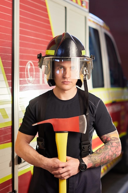 Foto de bombeiro usando capacete com machado contra carro de bombeiros Imagem de jovem bombeiro com capacete na cabeça contra o fundo do caminhão de bombeiros