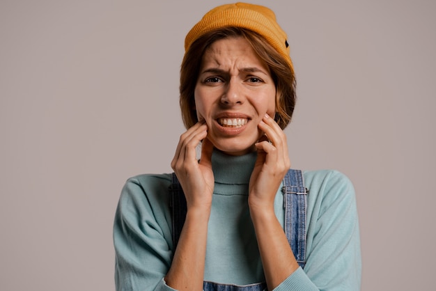 Foto foto de bonito hipster feminino segura o rosto e parece que ela tem dor nos dentes. mulher branca usa macacão jeans e fundo de cor cinza com chapéu isolado.