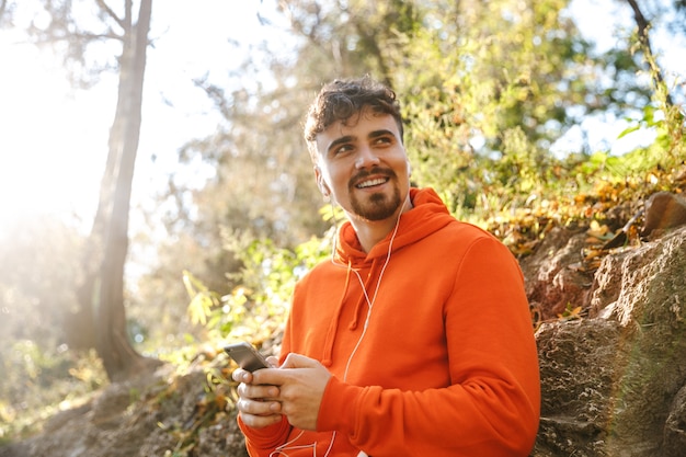 Foto de bonito jovem feliz esportes fitness homem corredor ao ar livre no parque, ouvindo música com fones de ouvido, usando telefone celular.