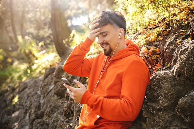 Foto de bonito jovem feliz esportes fitness homem corredor ao ar livre no parque, ouvindo música com fones de ouvido, usando telefone celular.
