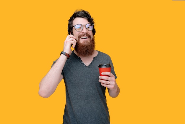 Foto de cara barbudo segurando um copo vermelho com café e falando no telefone amarelo isolado