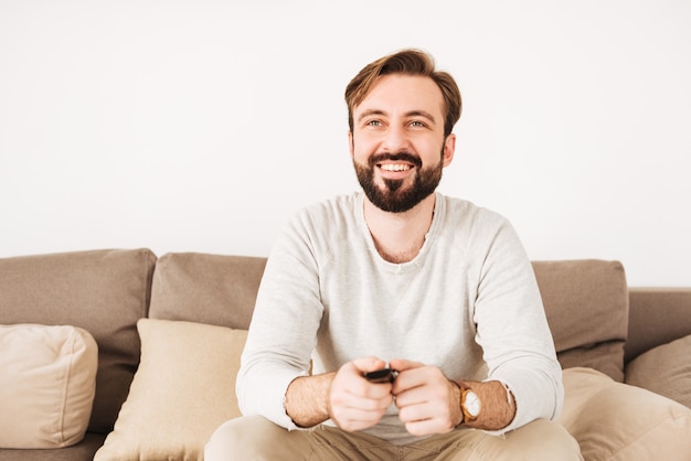 Foto de cara feliz, com barba e bigode, descansando em casa no sofá e assistindo TV com controle remoto nas mãos