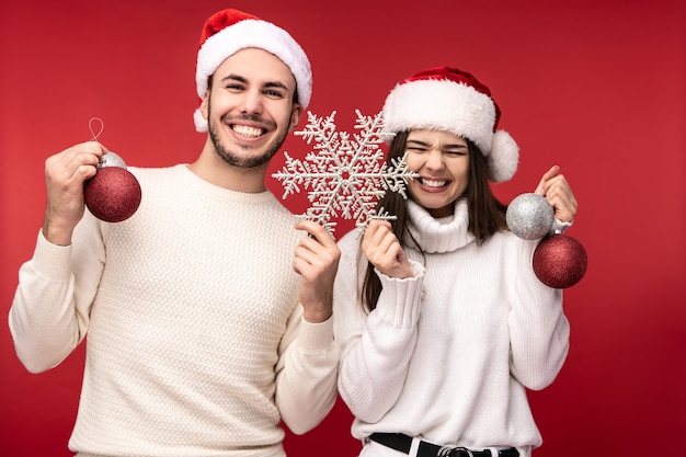 Foto foto de casal doce com chapéus de natal e brinquedos de natal, feliz com as férias. macho e fêmea estão apaixonados parecem abençoados e sorriem, isolados sobre fundo vermelho.