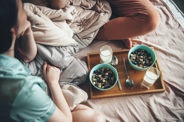 Foto de cima de um casal caucasiano comendo cereais com leite na cama, olhando um para o outro e curtindo o tempo juntos