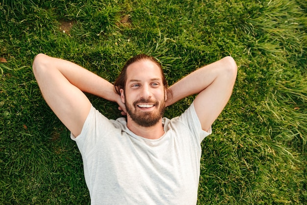 Foto de cima de um homem bonito barbudo de 30 anos, vestindo uma camiseta branca, deitado na grama em um parque verde, colocando as mãos atrás das costas