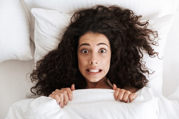 Foto de cima de uma divertida mulher tensa de 20 anos, com cabelo escuro encaracolado fazendo careta, deitada na cama sob um cobertor branco