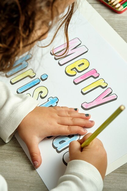 Foto de cima de uma garota irreconhecível colorindo letras de feliz natal com lápis de cor