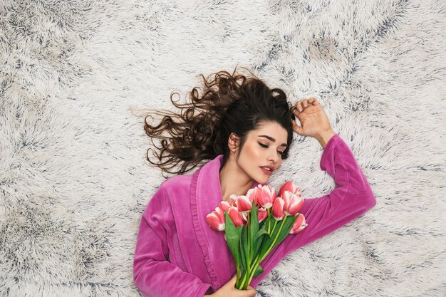 Foto de cima de uma mulher bonita com cabelo longo encaracolado, vestindo um roupão de menina, segurando flores enquanto está deitada sobre pelo branco no apartamento