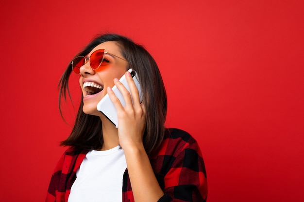 Foto de close de atraente morena jovem sorridente, vestindo uma camiseta branca elegante de camisa vermelha e óculos de sol vermelhos, isolados sobre um fundo vermelho, comunicando-se no celular, olhando para cima