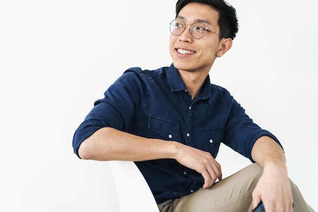 Foto de close de um homem asiático bonito de 30 anos usando óculos, sentado na cadeira e olhando para copyspace na luminosa sala de escritório