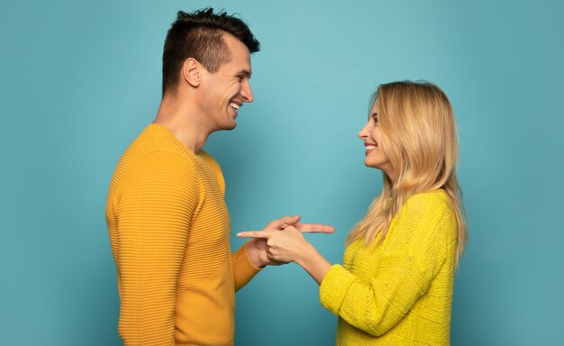 Foto de close de um lindo casal em suéteres amarelos, que está posando de perfil, olhando nos olhos um do outro com um sorriso e apontando um para o outro.