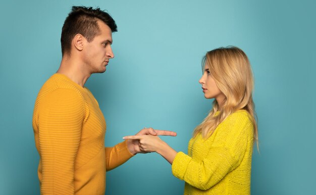 Foto de close de um lindo casal em suéteres amarelos, que está posando de perfil, olhando nos olhos um do outro com um sorriso e apontando um para o outro.