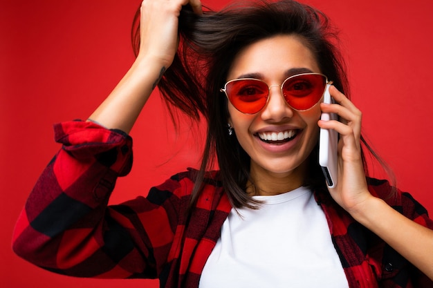 Foto de close de uma morena jovem sorridente e atraente, usando uma camiseta branca elegante e óculos de sol vermelhos