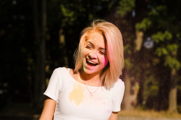 Foto de close de uma mulher loira alegre em uma camiseta branca com tinta colorida cobrindo o rosto no Festival de Holi