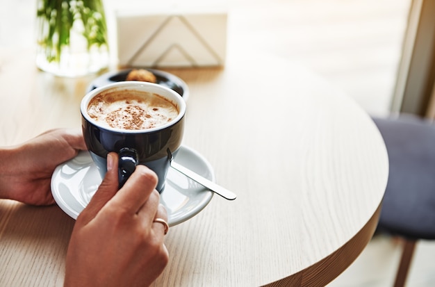 Foto de close-up de mãos femininas segurando uma xícara de cerâmica azul com cappuccino. Hora do café. Copie o espaço. Conceitos de comida e bebida