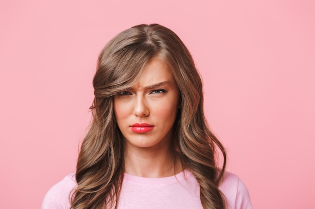 Foto de close up de mulher chateada com longos cabelos cacheados em uma camiseta básica fazendo beicinho e franzindo a testa em ressentimento, isolado sobre um fundo rosa