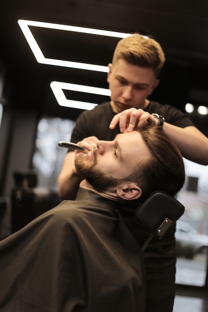 Foto de close-up de um cabeleireiro profissional, que está penteando a barba de sua jovem cliente, que se olha no espelho durante o procedimento.