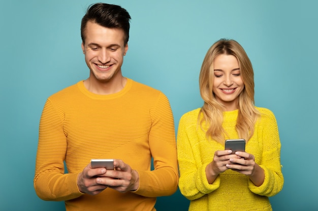 Foto de close-up de um casal de família em suéteres amarelos, em pé na frente e navegando na internet em seus smartphones.