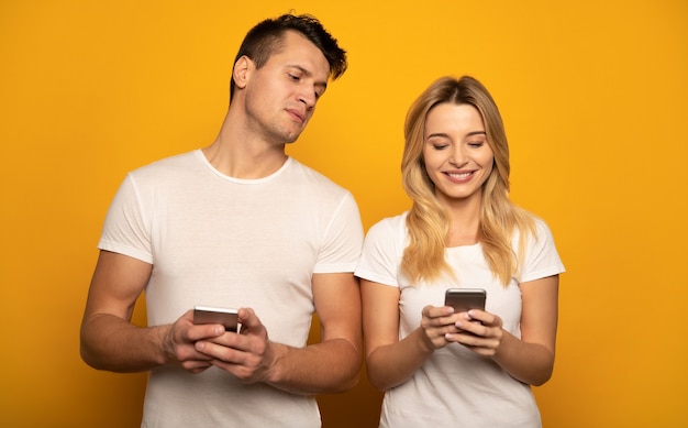 Foto de close-up de um casal fofo em camisetas brancas, que está posando na frente e navegando na internet em seus smartphones