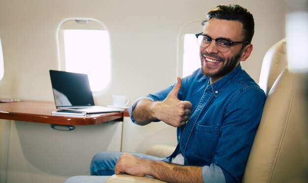 Foto de close-up de um homem bonito em uma roupa casual, que está sorrindo, enquanto digita algo em seu laptop e segura um smartphone na mão esquerda, voando na primeira classe de avião.