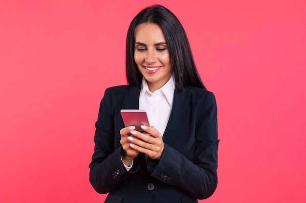 Foto de close-up de uma deslumbrante jovem morena de negócios com uma roupa elegante, que está segurando um smartphone nas mãos e sorrindo enquanto verifica o feed