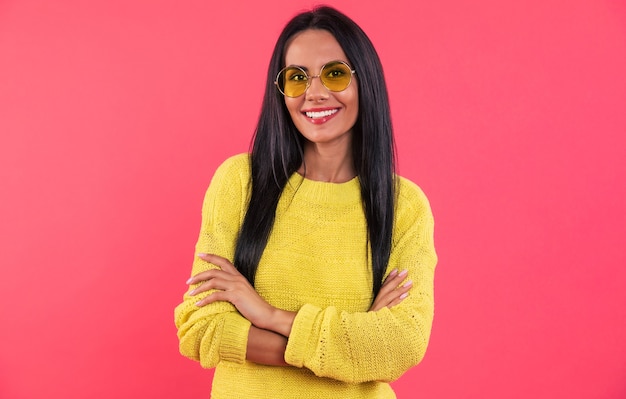 Foto de close-up de uma garota legal em um suéter de tricô amarelo e óculos de sol amarelos, que está posando com os braços cruzados e sorrindo para a câmera