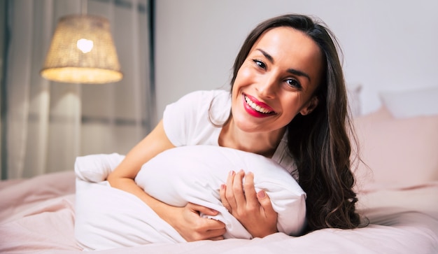 Foto de close-up de uma jovem magnífica, deitada na cama de bruços, abraçando um travesseiro e sorrindo sinceramente para a câmera.