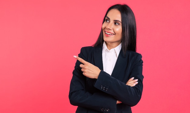 Foto de close-up de uma jovem mulher de negócios feliz, com longos cabelos negros, posando em um terno preto, olhando para a câmera e apontando para longe