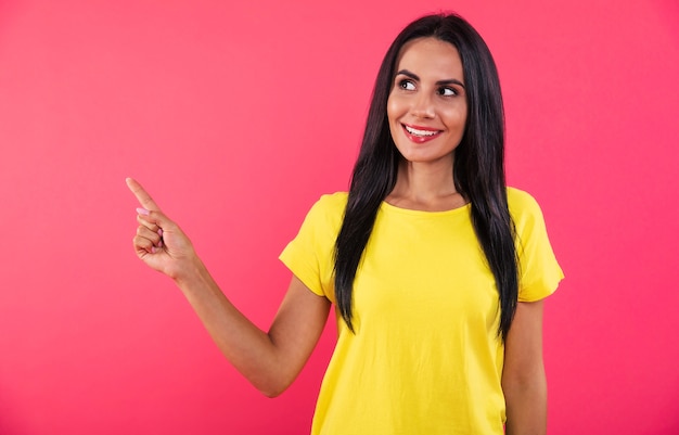 Foto de close-up de uma linda garota feliz, apontando para a esquerda com o dedo indicador direito e posando com uma camiseta amarela em um fundo rosa