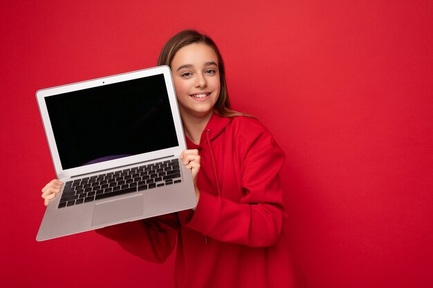 Foto de close-up de uma linda garota feliz e sorridente com cabelo comprido usando um capuz vermelho segurando um laptop