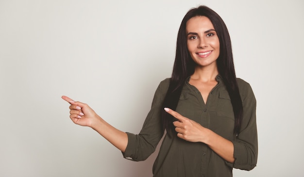 Foto de close-up de uma linda mulher em uma camisa cáqui, que está olhando para a câmera, sorrindo amplamente e apontando para a esquerda com o dedo indicador