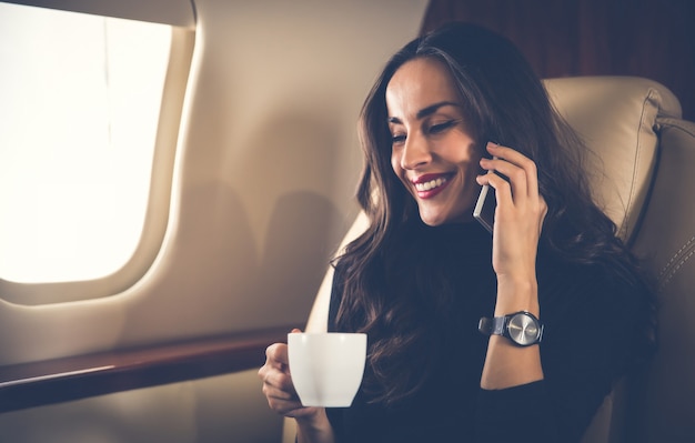 Foto de close-up de uma linda mulher em uma roupa casual, que está falando ao telefone e bebendo café preto durante seu voo em jatinho particular.