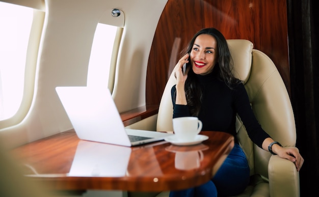 Foto foto de close-up de uma linda mulher em uma roupa casual, que está falando ao telefone e bebendo café preto durante seu voo em jatinho particular.